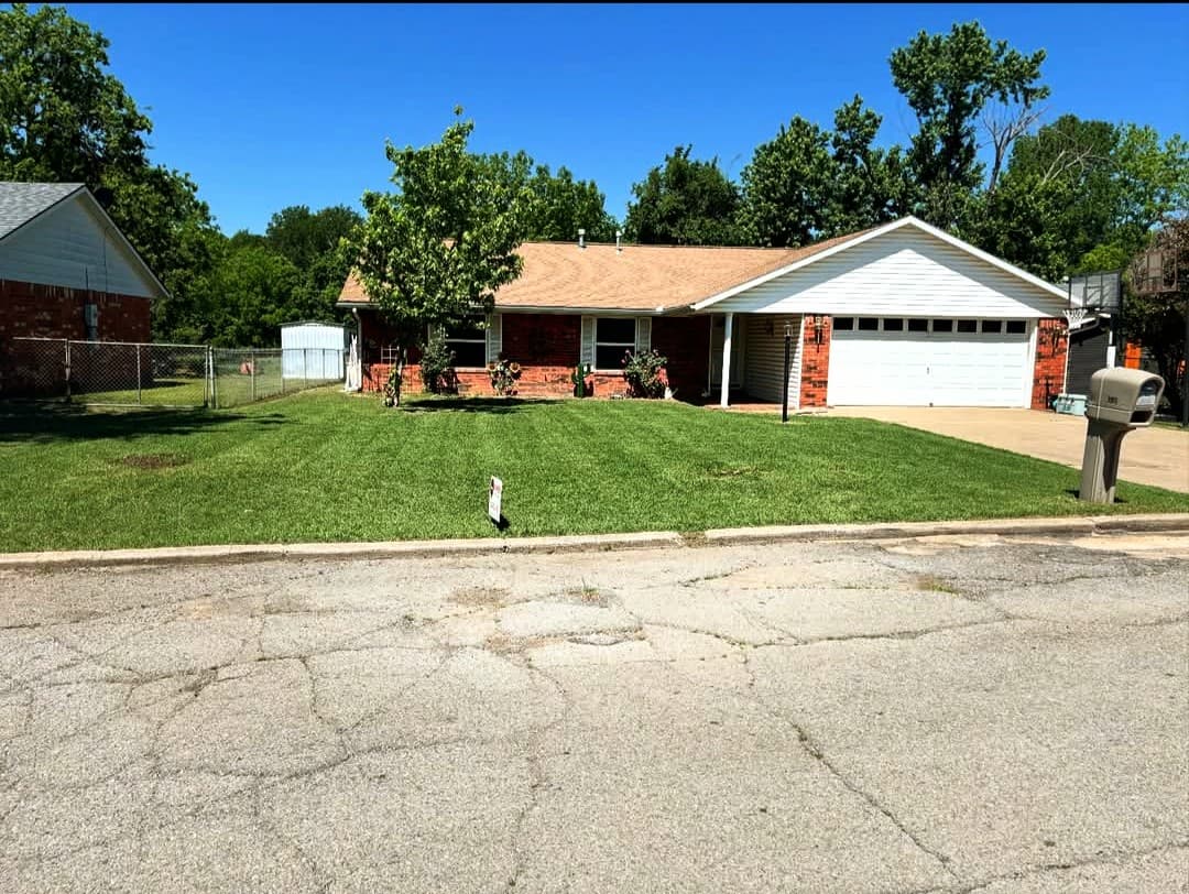 Red brick ranch house with a white garage door and green lawn on cracked road.