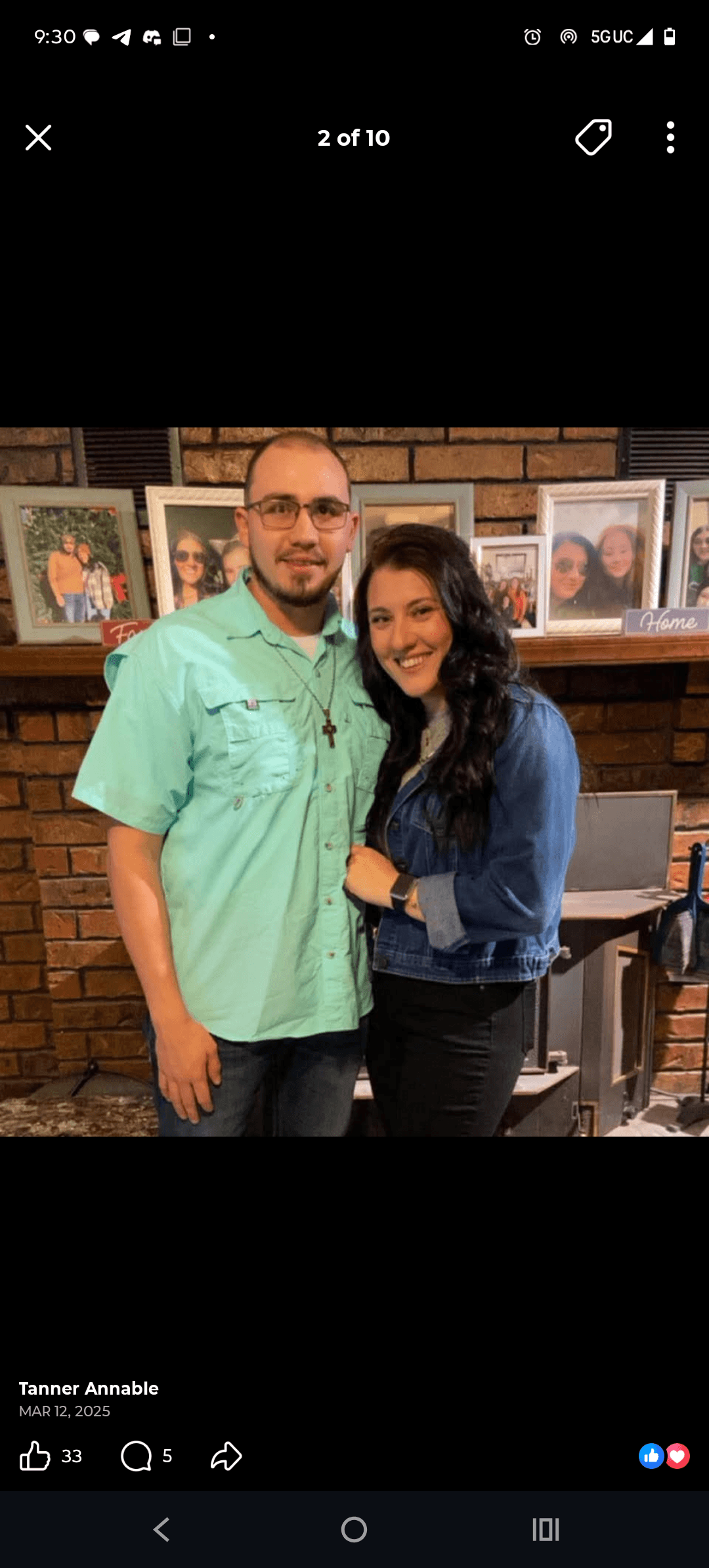 Smiling man and woman pose together in front of a brick fireplace with framed photos.