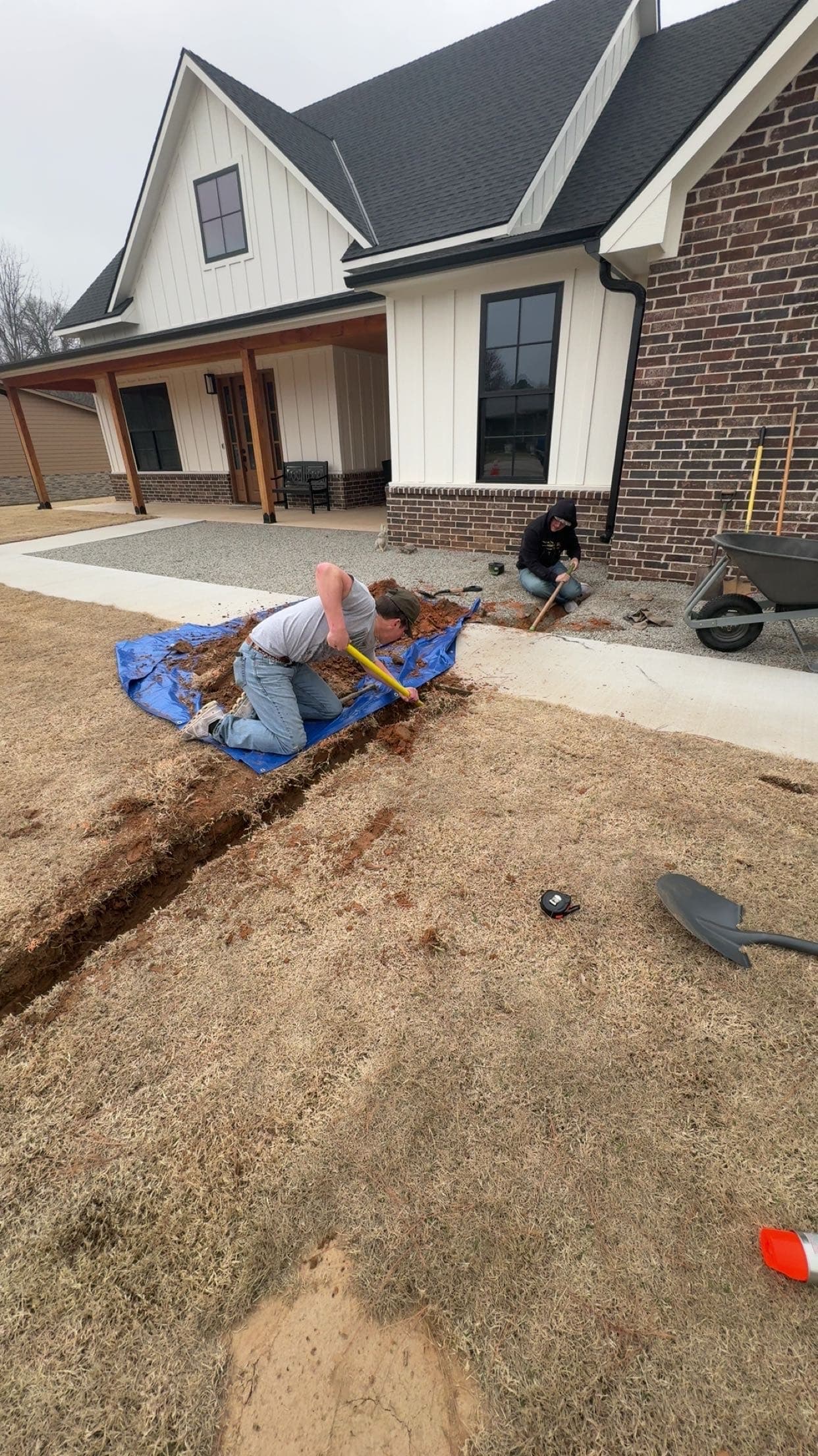 Two workers dig a trench in a dry lawn outside a modern white farmhouse.